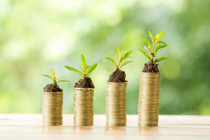 Münzen auf Holztisch vor grünem Hintergrund. Auf den Münzen kleine Pflanzen mit Erde als Symbol der Investition und des Sparens von Geld.